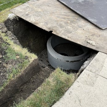 Goode Plumbing - Basin for sewer pump. - A concrete sewer pipe is partially exposed in a trench adjacent to a sidewalk, with the surrounding soil and grass visible. Two plywood sheets are placed over the trench, covering part of the excavation site. The trench appears to be part of ongoing plumbing or sewer work, with the pipe positioned below ground level and the area prepared for further construction or repair. - Plumbing Near Me - Chicago, Illinois