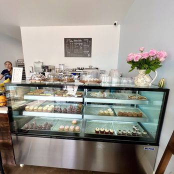 Dulce Cielo - Lots of homemade cupcakes! And the very nice owner Veronica! - A bakery display case showcases various cupcakes and desserts, with a menu board and a vase of pink roses on top. - Bakeries Near Me - Monrovia, California
