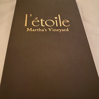 L'etoile Restaurant - A black, textured menu cover features gold lettering that reads 'l'étoile Martha's Vineyard,' indicating a restaurant located on Martha's Vineyard. The menu is placed on a white tablecloth, with a wine glass partially visible in the top left corner. The design suggests a formal dining setting. - French Near Me - Edgartown, Massachusetts