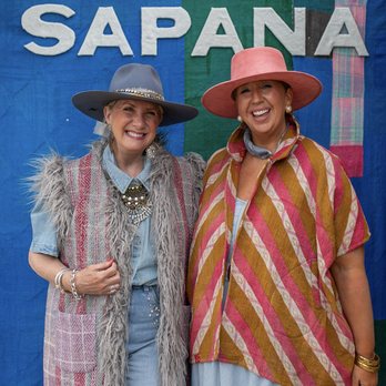 Sapana - Two women wearing wide-brimmed hats and vintage-inspired outfits stand in front of a backdrop with "SAPANA" displayed, smiling at the camera. - Women's Clothing Near Me - Round Top, Texas