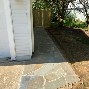 CM Hardscapes - Side walkway - A stone pathway curves toward a wooden gate in a backyard, bordered by a house wall and a garden with trees and grass. - Masonry/Concrete Near Me
