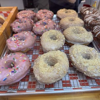Flatfish Gnv - Multiple donuts are arranged on a wire cooling rack, featuring pink-glazed donuts with multicolored sprinkles and white-glazed donuts with crumb topping. A person is seen in the background using tongs, indicating a bakery or donut shop environment. The donuts are displayed on a wooden tray with a red and white checkered paper underneath, suggesting a fresh, small-batch bakery setting. - Donuts Near Me - Gainesville, Florida