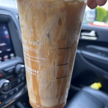 Starbucks - A large iced coffee beverage in a clear Starbucks cup with measurement markings is held by a person inside a vehicle. The drink contains milk and coffee, creating a marbled appearance. The background shows a car dashboard, steering wheel, and part of the windshield with some outdoor scenery. The cup features Starbucks branding and rewards information. - Coffee & Tea Near Me - Wappingers Falls, New York