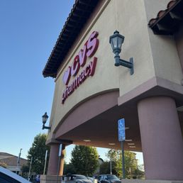 The image shows the exterior of a CVS Pharmacy store with a large sign displaying the CVS logo and the word 'pharmacy.' The building has a beige and pink facade with decorative lantern-style light fixtures. There is a parking lot in front of the store with several cars parked, including a visible white vehicle in the foreground. A blue handicap parking sign is also present near the entrance. The sky is clear and blue, indicating a sunny day, and some trees are visible in the background.