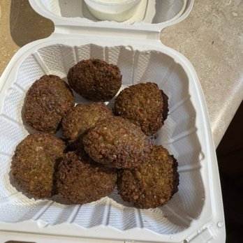 Shalimar - Falafel (Vegan) - The image shows a white takeout container filled with multiple fried falafel balls, which are round and golden-brown with a crispy exterior. A small plastic cup of creamy white sauce, possibly tahini or yogurt-based, is placed in the lid of the container. The container is on a textured surface, and the setup suggests a meal from a Middle Eastern or Mediterranean cuisine restaurant. - Indian Near Me - Delmar, New York