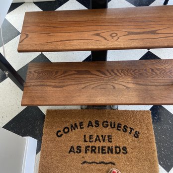 Artizen - Cute mat - A rectangular doormat with the text "Come as guests, leave as friends" in black letters on a brown background is positioned on a black and white checkered floor. The doormat is in front of two wooden benches with a natural finish, supported by black metal frames. A person's foot with painted toenails and a sandal is visible at the bottom of the image. - Coffee & Tea Near Me - The Bronx, New York