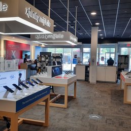 Inside the store - The photo depicts the interior of an Xfinity Store by Comcast, with display tables showcasing mobile phones and accessories. Customer service counters are visible, with staff assisting customers. The store has a modern design with a dark ceiling, bright lighting, and large windows at the front allowing natural light. Signage with the Xfinity Mobile branding is prominently displayed overhead. The space appears organized and ready for customer interactions.