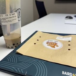 Brown sugar boba and empty bubble waffle wrapper - A bubble tea drink with brown sugar tapioca pearls and a straw is positioned on a table. Next to it is a paper bag featuring a Boba King sticker with a cartoon character. The table also holds a black folder or menu. In the background, a monitor and a notebook are visible, indicating an indoor setting, possibly an office or meeting room.