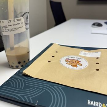 Boba King - Brown sugar boba and empty bubble waffle wrapper - A bubble tea drink with brown sugar tapioca pearls and a straw is positioned on a table. Next to it is a paper bag featuring a Boba King sticker with a cartoon character. The table also holds a black folder or menu. In the background, a monitor and a notebook are visible, indicating an indoor setting, possibly an office or meeting room. - Bubble Tea Near Me - Wheaton, Illinois