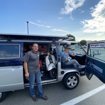 Premier Mobile Locksmith - The image shows a man standing outside a small, dark-colored van with an open sliding door, revealing a scooter inside. A woman is seated in the driver's seat, wearing a hat, sunglasses, and casual clothing, holding a drink and looking at the camera. The parking lot is surrounded by other vehicles, and the sky above is partly cloudy with patches of blue. The scene appears to be taken during daylight hours, with a clear and bright atmosphere. - Keys & Locksmiths Near Me - Tustin, California