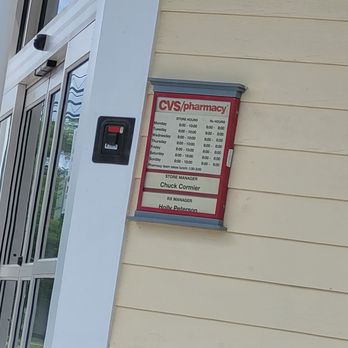 CVS Pharmacy - Hours - A store hours sign for CVS pharmacy is attached to a beige horizontal siding wall outside the store. The sign displays the weekly hours for each day and the lunch break times for the pharmacy team. The sign is framed with a gray border and positioned near a glass door with metal framing, which is partially visible on the left side of the image. - Drugstores Near Me - Canton, Connecticut