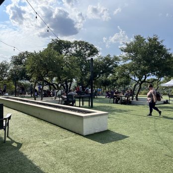Frontyard Brewing - Spacious outdoor area with trees, tables, and chairs at Frontyard Brewing in Spicewood, TX. People are seated and walking, with string lights hanging overhead. The sky is partly cloudy, and there is a white canopy tent in the background. The setting includes a grassy surface and shaded seating options, suitable for social gatherings and outdoor activities. - Breweries Near Me - Spicewood, Texas