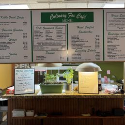 The menu - The image shows a large, illuminated menu board at Culinary Fox Cafe in Elgin, IL, featuring sections for sandwiches, salads, soups, and beverages. The menu includes items like chicken meatball romano, focaccia grinders, and specialty salads. Below the board, a counter holds potted plants, a handwritten sign with meal deals, and some kitchen equipment. The setting appears to be a casual cafe interior with a focus on diverse, handcrafted food offerings.