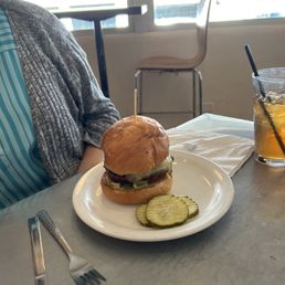 A burger with lettuce, tomato, and other toppings on a white plate, with sliced pickles, served in a casual dining setting with a glass of iced tea.