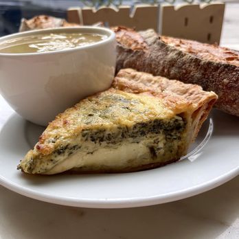 Verzenay - Kale and Leek Quiche, Asparagus Soup, Baguette - A plate holds a slice of quiche with spinach and cheese, a bowl of soup, and a baguette bread, set near a window. - Desserts Near Me - Chicago, Illinois