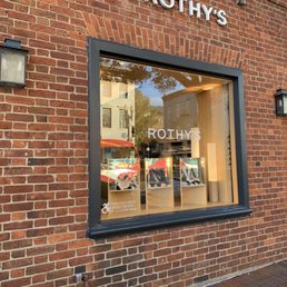 The image shows the exterior of Rothy's, a shoe store, with a brick facade and a large display window. Inside the window, shoes are arranged on stands, and the store's name, Rothy's, is displayed on the glass. The window reflects nearby buildings and trees. There are exterior wall-mounted lights on either side of the window, and a small sign indicating accessibility options is visible at the bottom left of the window. The sidewalk in front is made of brick pavers.