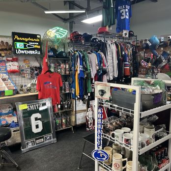 Simpler Times - Sports jerseys, and collectibles. - The store contains sports jerseys, helmets, and collectibles displayed on racks and shelves. There are vintage sports memorabilia, including framed jerseys and helmets, along with mugs and other memorabilia on a white wire rack. The background features a pegboard with various sports items, a neon sign, and a mix of sports apparel and accessories. The space appears cluttered but organized for browsing sports collectibles. - Antiques Near Me - Palm, Pennsylvania