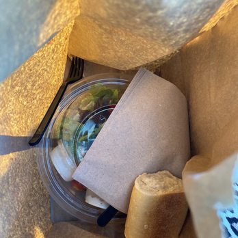 Panera Bread - The image shows the interior of a paper bag with a salad in a clear plastic container, a bread roll, and a black plastic fork. The salad appears to have greens and possibly other vegetables. The bread roll is partially eaten or broken. The items are arranged inside the bag, indicating a takeout or packed meal. - Sandwiches Near Me - Ridgeland, Mississippi