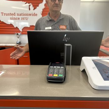 Public Storage - A man with glasses and a gray shirt stands behind a checkout counter at Public Storage, with a computer monitor, payment terminal, and a white device on the counter. - Self Storage Near Me - Glendale, Arizona