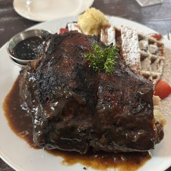 Flamingobaby Kitchen - Jerk Chicken & Waffles - A plate features braised meat with dark sauce, mashed potatoes, waffles dusted with powdered sugar, and a small container of syrup, served on a white dish. - Caribbean Near Me - Brooklyn, New York