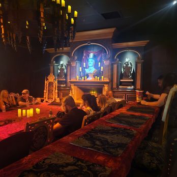 Mystique The Magic Experience - Guests sit at a long, red-covered table in a dark room with theatrical decor, including a fireplace, statues, and candles, at Mystique The Magic Experience. - Magicians Near Me - Mesa, Arizona