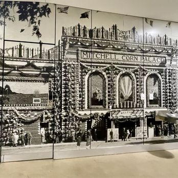 Corn Palace - A large black-and-white mural shows the facade of the Mitchell Corn Palace, including its ornate architectural details and signage. The mural features people walking along the sidewalk in front of the building, with some standing near the entrance. The scene captures the historic and decorative style of the Corn Palace, emphasizing its significance as a landmark in Mitchell, South Dakota. - Museums Near Me - Mitchell, South Dakota