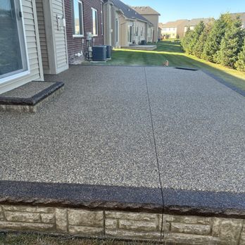 Unique Cement - The image shows a textured concrete patio attached to a residential building, bordered by a stone foundation. A small step with a textured surface is present near the house, and a drain is visible on the patio surface. The background features a grassy yard with trees and neighboring houses, indicating a suburban setting. - Masonry/Concrete Near Me - Clinton Township, Michigan