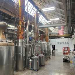 The image depicts the interior of KOVAL Distillery in Chicago, showcasing large copper and stainless steel distillation stills and equipment. The space has a high, angled ceiling with skylights allowing natural light, and fluorescent lighting fixtures. An American flag hangs in the background, and the distillery's name is visible on the wall. The floor is concrete, and there are various tools and a small lift present, indicating an industrial, production environment.