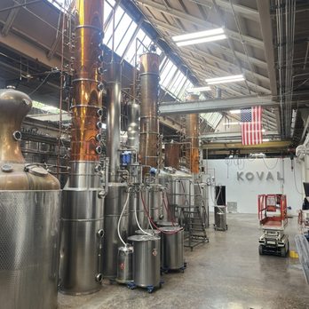 KOVAL Distillery - The image depicts the interior of KOVAL Distillery in Chicago, showcasing large copper and stainless steel distillation stills and equipment. The space has a high, angled ceiling with skylights allowing natural light, and fluorescent lighting fixtures. An American flag hangs in the background, and the distillery's name is visible on the wall. The floor is concrete, and there are various tools and a small lift present, indicating an industrial, production environment. - Distilleries Near Me - Chicago, Illinois