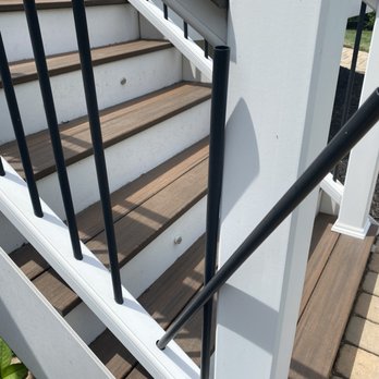 Sunburst Construction - Poles won't stay in handrails - A staircase with wooden treads and white risers, featuring black metal balusters. Two sections of the handrail are missing, with one section leaning against the wall and another detached. The staircase is outdoors, adjacent to a brick-paved walkway and a landscaped area with mulch and grass. - General Contractors Near Me - Leesburg, Virginia