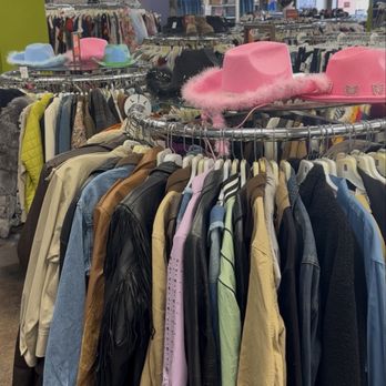 Buffalo Exchange - The image shows a thrift store interior with multiple circular clothing racks filled with jackets, shirts, and other apparel in various colors and styles. Several pink hats with feather trim are placed on top of the racks. The background reveals more clothing displays, shelves, and store fixtures, with a spacious layout and fluorescent lighting. - Women's Clothing Near Me - Austin, Texas