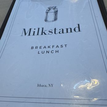 Milkstand - Menu - A menu from Milkstand in Ithaca, NY, is shown with a minimalistic design, featuring a jar illustration at the top. The menu lists breakfast and lunch options and is encased in a black cover. The background appears to be a light-colored table surface. - Breakfast & Brunch Near Me - Ithaca, New York