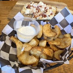 Urban Spicy Fries- maybe I missed out on the spicy part!? - A basket filled with fried potato wedges, seasoned and crispy, is lined with black and white checkered paper and accompanied by a small cup of creamy dipping sauce. In the background, a pizza slice with toppings such as cheese and possibly bacon or ham is on a wooden serving board. The setting appears to be a casual dining environment, likely a restaurant or pizzeria.