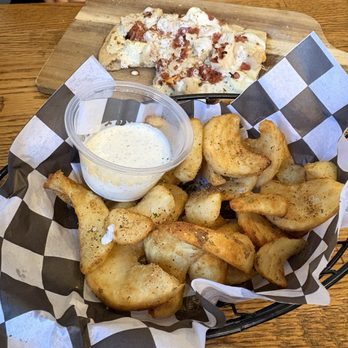 Urban Pine Eatery - Urban Spicy Fries- maybe I missed out on the spicy part!? - A basket filled with fried potato wedges, seasoned and crispy, is lined with black and white checkered paper and accompanied by a small cup of creamy dipping sauce. In the background, a pizza slice with toppings such as cheese and possibly bacon or ham is on a wooden serving board. The setting appears to be a casual dining environment, likely a restaurant or pizzeria. - Pizza Near Me - Flagstaff, Arizona