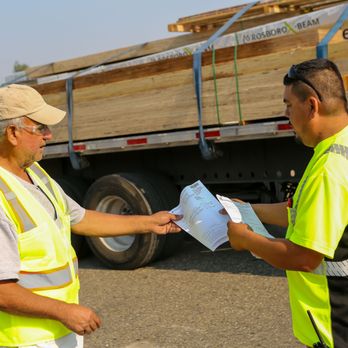 Builders FirstSource - Two Builders FirstSource employees wearing high-visibility safety vests are reviewing paperwork in front of a flatbed truck l... - Building Supplies Near Me - Petoskey, Michigan