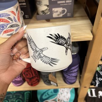 Mestiza - Some of the mug varieties they had, and reasonably priced. - A ceramic mug featuring black and white eagle and owl illustrations is held by a person with decorated nails, with shelves of other mugs and items in the background. - Jewelry Near Me - Chicago, Illinois