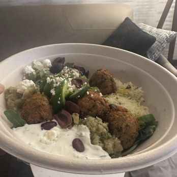 CAVA - Greens & grains bowl $15.99 missing ingredients & not even half full. Left out my pita, too. 2 leaves of arugula. RIP OFF - A bowl of Mediterranean cuisine containing falafel, vegetables, and toppings is held in front of a sofa with pillows in the background. - Mediterranean Near Me - Brunswick, New Jersey