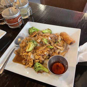 Chilli Thai - Pad See ew - A square white plate contains Pad See Ew with stir-fried noodles, broccoli, and pieces of meat, accompanied by a spring roll and a small cup of dipping sauce. The dish is served on a dark wooden table, with a spoon placed on the left side of the plate. In the background, there are condiment containers with sauces and a wooden chair. The setting appears to be inside a restaurant, likely an Asian or Thai cuisine establishment. - Thai Near Me - Carrollton, Texas