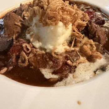 636 Main Ave - Red wine Beef Tips roasted mashed potatoes - A close-up of a bowl containing beef stew with visible chunks of beef and vegetables in a dark broth. The dish is topped with a dollop of mashed potatoes and crispy fried onions. The presentation suggests a hearty, savory meal served in a restaurant environment. - New American Near Me - Durango, Colorado