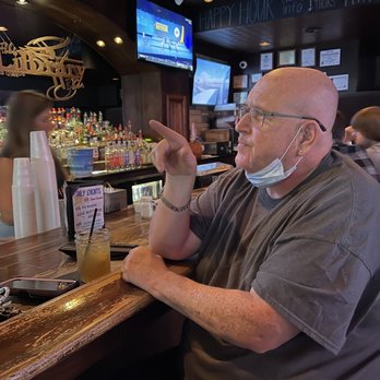 Library Bar - a man wearing a face mask - A man with glasses and a face mask hanging around his neck is sitting at a bar counter, pointing with his right hand. He has a shaved head and is wearing a gray T-shirt. The bar has bottles of alcohol, cups, and a drink in front of him. Behind the bar, there are two television screens and a chalkboard sign with happy hour information. Other patrons are visible in the background, and the setting appears to be a casual bar or pub. - Beer Bar Near Me - Bourbonnais, Illinois
