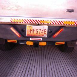 The photograph captures the rear of a vehicle with a New Mexico license plate HFG748, mounted on a black bumper with reflective red and yellow safety markings. The vehicle has a trailer hitch and a visible exhaust pipe on the right side. The background includes part of another vehicle with a Ford emblem and a textured mat surface beneath the vehicle. The image appears to be taken indoors or in a garage setting.