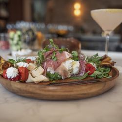 The Farm Italy - Antipasti platter with prosciutto, salami, burrata, cheese, roasted tomatoes, greens, and vegetables served on a wooden platter.
