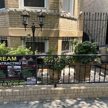 Dream Contracting - Power Washing - A front porch area features large potted plants, a black metal railing, and a sign advertising Dream Contracting Inc. The background shows a brick building with windows and a gated entrance. - Masonry/Concrete Near Me - Brooklyn, New York