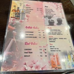 This menu displays a selection of Japanese beverages including beers like Sapporo, Kirin Ichiban, Heineken, and Corona, with prices for small and large sizes. It also offers white wines (Chardonnay, Pinot Grigio, Sauvignon Blanc) and red wines (Cabernet, Pinot Noir, Merlot), all priced per glass and bottle. The sake options include house hot and cold sake, and various flavored sake, with prices listed. Soft drinks such as Ramune soda, Coke, Fanta, and ginger ale are available, along with juices and teas. The design features cherry blossom graphics, creating a floral theme.