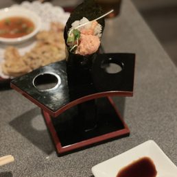 Spicy Tuna Rolls - A sushi handroll with fish, rice, and greens is displayed on a black stand, with a small dish of soy sauce in front.