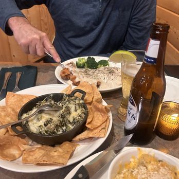 149 Eatery - Chicken fried chicken and spinach dip - A person wearing a dark gray zip-up jacket is seated at a wooden table in a restaurant, cutting into a plate of spinach dip topped with black pepper, accompanied by broccoli. On the table, there is a bowl of baked spinach with cheese, a bottle of beer, a glass of beer with a lemon wedge, and a small candle holder. The setting appears cozy with wooden paneling in the background. - American Near Me - Creede, Colorado