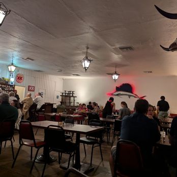 El Torero - The restaurant interior features several tables and chairs with patrons seated and dining. The ceiling has hanging lantern-style lights, and the walls are decorated with a mounted bull head on the right and a large fish with a red outline on the back wall. The bar area on the left has bottles and signs, including a neon sign. The overall setting suggests a casual dining environment with themed decor. - Mexican Near Me - Tucson, Arizona