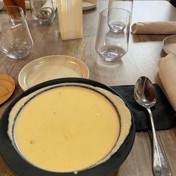 World Spa - Lobster bisque - A bowl of lobster bisque is placed on a wooden dining table, surrounded by empty glasses, a napkin, and small plates, with a window in the background. - Day Spas Near Me - Brooklyn, New York