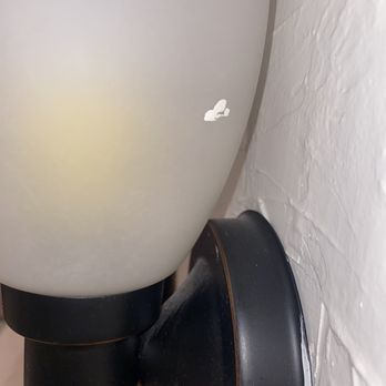 Happy Cleaning LA - Wall sconce with dust after "deep cleaning" - A wall sconce with dust is attached to a white textured wall, with a small butterfly sticker nearby. The sconce has a black base and a white shade. - Home Cleaning Near Me - Glendale, California