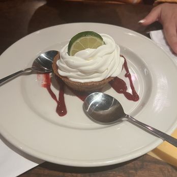 The Tombs - Key lime pie was the perfect dessert after our shrimp and grits and crab cake! - A key lime pie dessert is presented on a white plate, topped with a swirl of whipped cream and garnished with a lime slice. The plate includes decorative drizzles of chocolate sauce and two spoons placed on either side of the dessert. The setting appears to be in a restaurant environment, with a dark wooden table and a person’s hand visible in the background. - New American Near Me - Washington, DC, District of Columbia